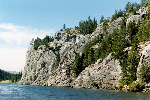 Rocky cliffs lined with evergreen trees rise above a calm river under a clear blue sky.