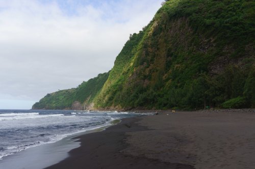 A serene beach with dark sand, gentle waves, and lush green cliffs under a partly cloudy sky.