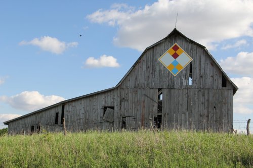A weathered barn with a colorful diamond pattern on its gable, surrounded by tall grass and a blue sky with clouds.