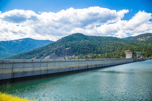 A concrete dam stretches across a serene lake, surrounded by lush green mountains and a blue sky with fluffy clouds.