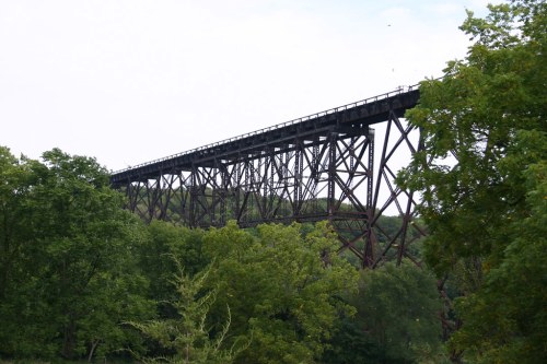 A tall, rusted railway trestle bridge spans over lush green trees under a cloudy sky.