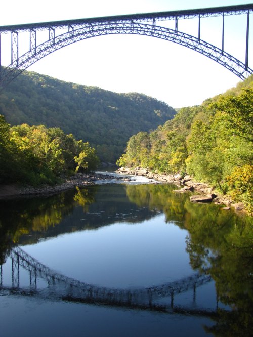A scenic view of a river reflecting a large bridge, surrounded by lush green hills under a clear sky.