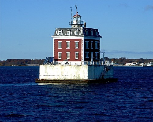 A red and white lighthouse stands on a small island in the water, surrounded by calm blue waves and a clear sky.
