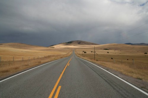 A long, empty road stretches through golden fields under a cloudy sky, leading towards distant hills.