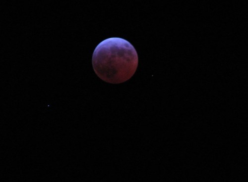 A large, reddish moon against a dark sky, with a few faint stars visible nearby.