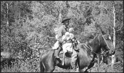 A man on horseback carries two children, surrounded by trees and greenery in a vintage black-and-white photo.