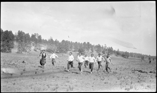 A group of eight men running in a field, surrounded by trees and a clear sky, in a vintage black-and-white photograph.
