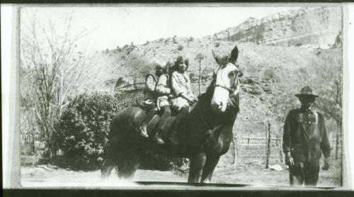 Two children sit on a horse, while a man stands nearby in a rural setting with hills in the background.