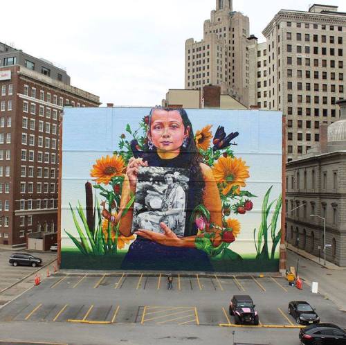 A vibrant mural of a girl holding a black-and-white photo, surrounded by colorful flowers and greenery on a city building.