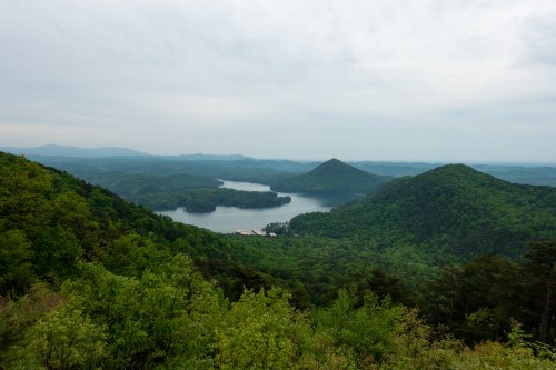 A panoramic view of lush green mountains and a serene lake under a cloudy sky.
