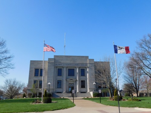 Henry County Courthouse with American and Iowa flags, surrounded by trees and a clear blue sky.
