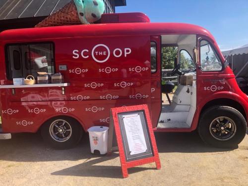 A red ice cream truck named "The Scoop" with a menu sign outside, parked in a sunny outdoor area.
