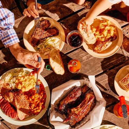 A table spread with various barbecue dishes, including ribs, steak, sides, and drinks, enjoyed by multiple people.