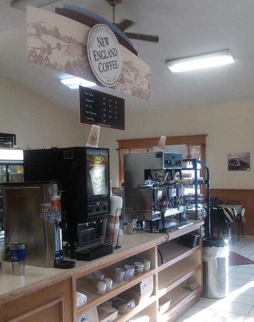A cozy coffee shop interior featuring a menu board, coffee machines, and a display of cups and beverages.