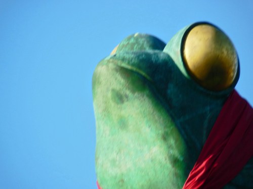 A large green frog statue with prominent golden eyes, wearing a red scarf, against a clear blue sky.