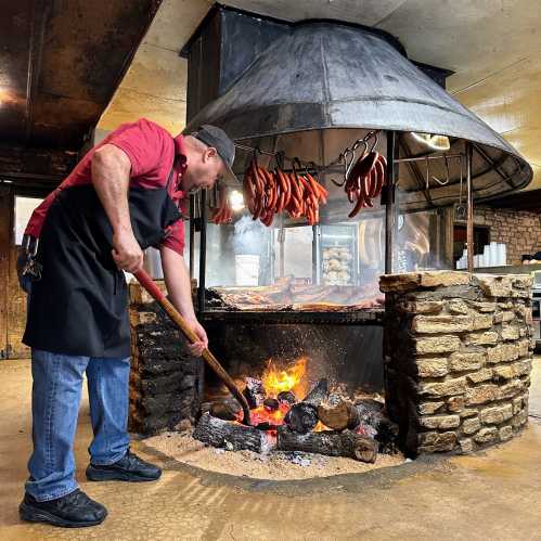 A chef stirs a fire in a stone pit, surrounded by hanging sausages and a grill in a rustic kitchen setting.