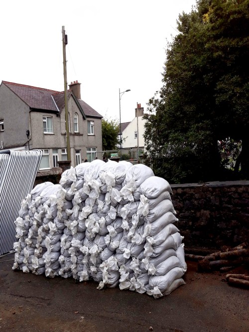 A large stack of white bags piled on a gravel surface, with houses and trees in the background.