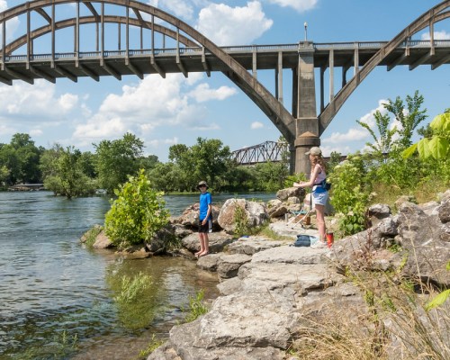 Two children stand on rocky riverbank under a large bridge, enjoying a sunny day by the water. Lush greenery surrounds them.