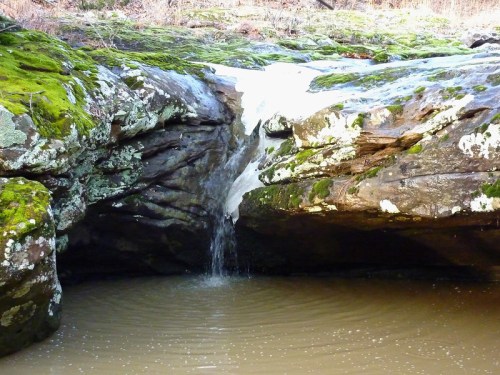 A small waterfall cascades over moss-covered rocks into a calm pool of water below.