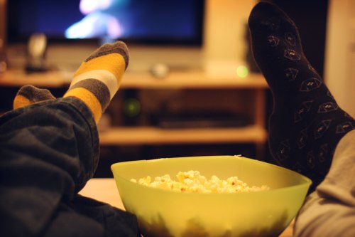 Two pairs of feet in colorful socks resting near a bowl of popcorn, with a TV in the background.