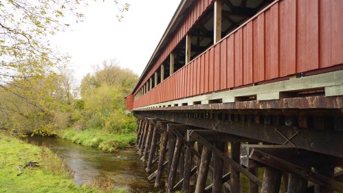 A red covered bridge stands on wooden supports over a small stream, surrounded by greenery and trees.