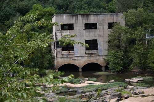 Abandoned concrete building with broken windows, surrounded by greenery and a small body of water.