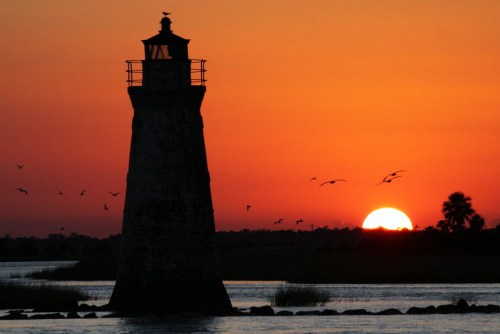 Silhouette of a lighthouse against a vibrant sunset, with birds flying and water reflecting warm colors.