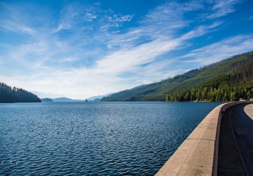 A serene lake surrounded by mountains and trees under a clear blue sky, with a curved concrete path along the water's edge.