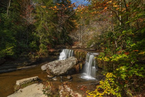 A serene waterfall cascades over rocks, surrounded by vibrant autumn foliage and lush greenery.