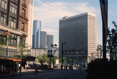 A bustling city scene with tall buildings, people walking, and trees lining the street under a clear blue sky.