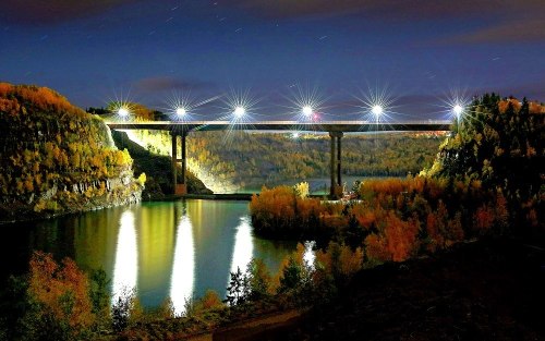 A brightly lit bridge spans a calm river, surrounded by autumn foliage under a starry night sky.