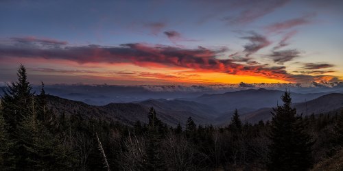 A stunning sunset over layered mountains, with vibrant clouds and a colorful sky, framed by evergreen trees.