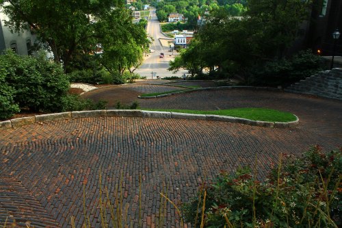A winding brick road descends through greenery, leading to a view of a town in the distance.