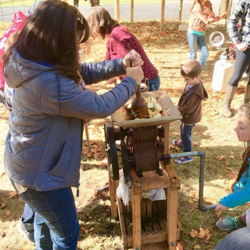 A person operates an apple press while children and others watch, surrounded by autumn leaves and a farm setting.