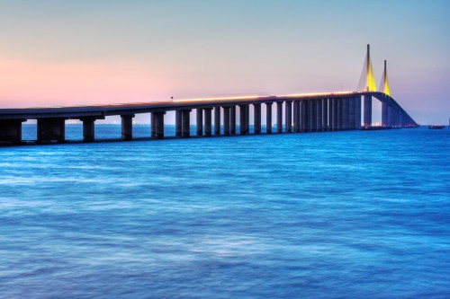 A long bridge spans over calm blue waters at dusk, with lights illuminating its structure against a colorful sky.