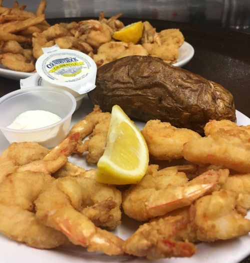 A plate of fried shrimp with a baked potato, lemon wedges, and dipping sauces, alongside a second plate of shrimp and fries.