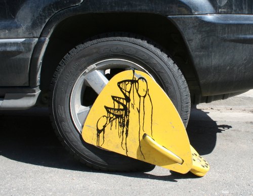 A yellow wheel clamp attached to a tire of a black vehicle, with graffiti-like drips on its surface.