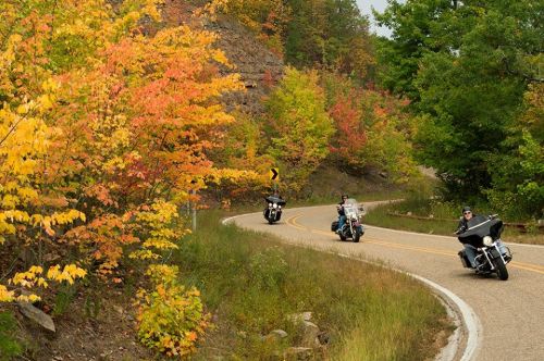Motorcyclists ride along a winding road surrounded by vibrant autumn foliage in shades of orange and green.