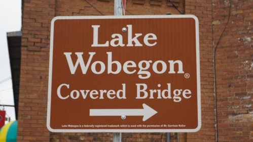 Sign for Lake Wobegon Covered Bridge with an arrow pointing to the right, set against a brick background.