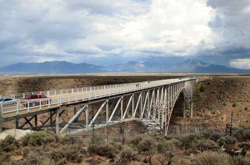 A long, elevated bridge spans a canyon, with mountains in the background and a cloudy sky above.