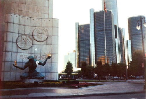A statue of a man holding a flame, with modern skyscrapers in the background and trees lining the foreground.