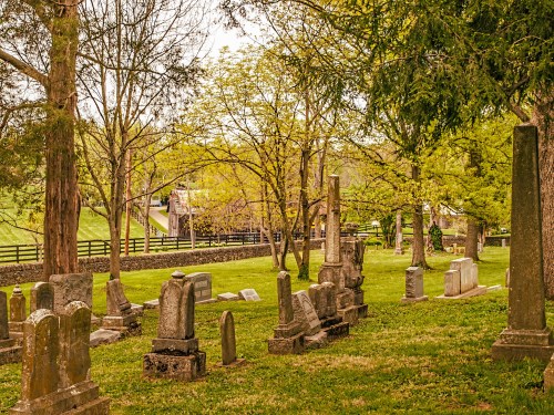 A peaceful cemetery with various gravestones surrounded by trees and greenery, set against a rural backdrop.