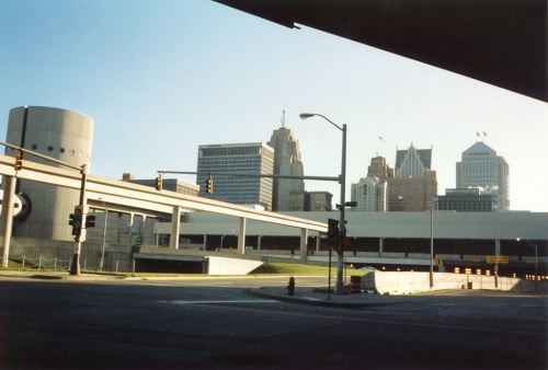 City skyline featuring tall buildings and overpasses under a clear blue sky. Traffic lights and street signs visible.