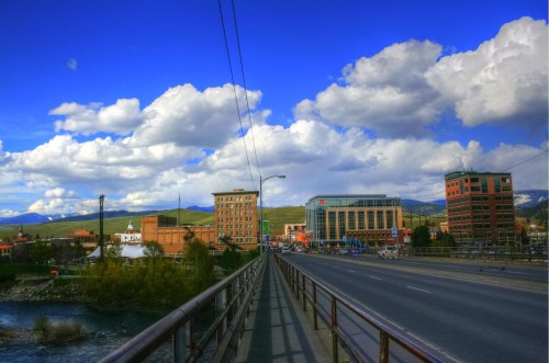 A bridge leads to a cityscape with modern and historic buildings under a blue sky with fluffy clouds.