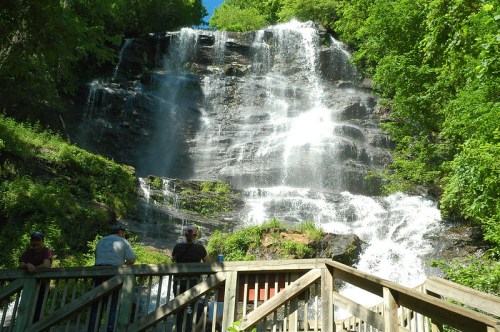 A scenic waterfall cascades down rocky cliffs, surrounded by lush greenery, with visitors enjoying the view from a wooden deck.