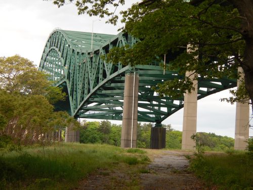 A green arch bridge spans over a grassy area, supported by concrete pillars, surrounded by trees and a cloudy sky.