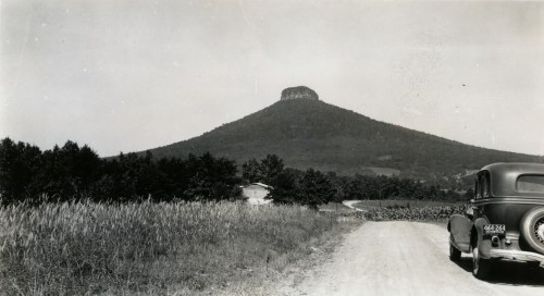 A vintage car on a dirt road with a mountain in the background, surrounded by trees and fields.