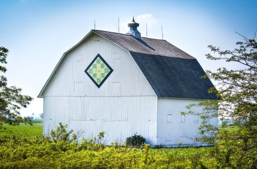 A white barn with a diamond-shaped mural, surrounded by green fields and trees under a clear blue sky.