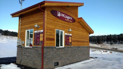 A small wooden restaurant with a stone base, featuring a menu board and snowy surroundings.