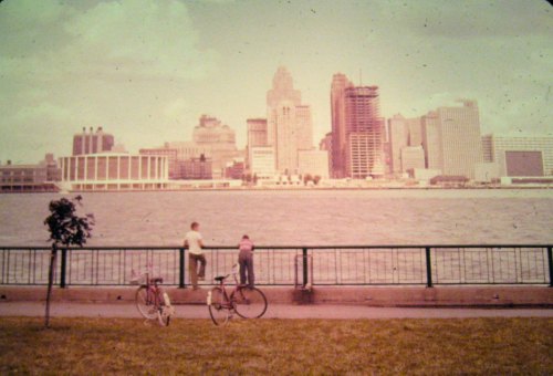 Two people lean on a railing by the water, with bicycles nearby and a city skyline in the background.